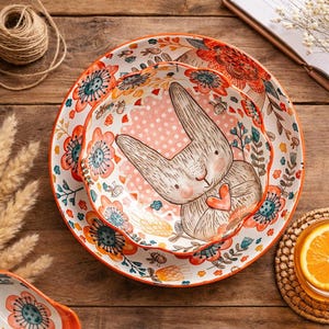 May include: Two ceramic plates with a floral and bunny design. The plates have a white background with orange, teal, and pink floral patterns. The centre plate features a bunny illustration with a heart.