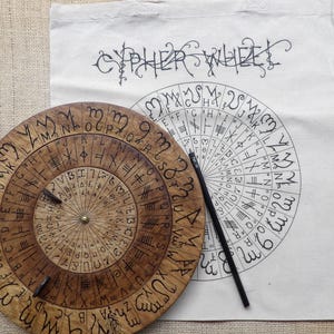 Può includere: Una ruota cifrata in legno con simboli incisi e un centro in ottone, accanto a una borsa di tela bianca con le parole "CYPHER WHEEL" e un disegno a ruota cifrata abbinato. Una matita nera poggia sulla tela.
