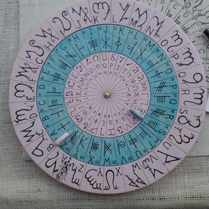 Könnte beinhalten: Ein rundes Runenrad aus Holz mit rosa und blauem Hintergrund. Das Rad hat ein drehbares Zifferblatt mit Runen und Buchstaben am Rand. Das Rad wird für Divination und Magie verwendet.