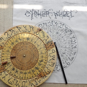 Peut inclure: Une roue de chiffrement en bois avec des symboles et des lettres gravés, à côté d'une toile imprimée avec les mots "CYPHER WHEEL" et un motif circulaire similaire. Un stylo noir repose sur la toile.