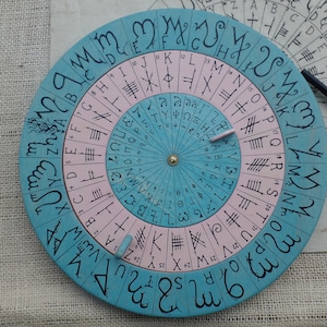 Peut inclure: Une roue d'alphabet en bois circulaire avec un schéma de couleurs bleu et rose. La roue a un cadran rotatif avec des lettres et des symboles autour du bord. La roue est sur une surface en toile de jute.