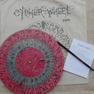 Könnte beinhalten: Ein hölzernes Chiffrierrad mit rotem und grauem Design. Das Rad hat einen drehbaren inneren Kreis mit Buchstaben und Symbolen. Das Rad befindet sich auf einer beigefarbenen Stoffoberfläche mit einem Bleistift und einem Blatt Papier mit dem Text "cipherwheel.com".