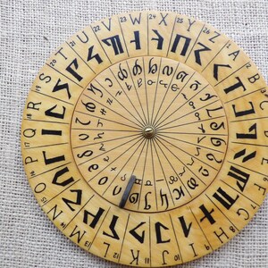 Może przedstawiać: Drewniane koło szyfrujące z czarnymi symbolami i literami ułożonymi w koncentryczne okręgi. Zewnętrzny pierścień zawiera litery i cyfry, podczas gdy wewnętrzny pierścień ma inny zestaw symboli. Metalowy wskaźnik jest umieszczony na kole.