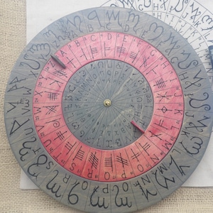 Puede incluir: Una rueda de runas circular de madera con un esquema de color rojo y gris. La rueda tiene un eje central con dos punteros. El anillo exterior está etiquetado con las letras del alfabeto, y el anillo interior está etiquetado con runas.