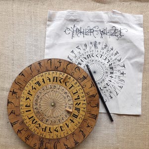 May include: A wooden cipher wheel with rotating inner dials, featuring engraved symbols and numbers. A black pen rests beside the wheel, and a canvas tote bag with the words "CYPHER WHEEL" is in the background.