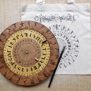 May include: A wooden cipher wheel with engraved symbols and letters, next to a white tote bag with the words "CYPHER WHEEL" and a matching cipher design. A black stylus rests nearby. The wheel is approximately 25 cm in diameter.