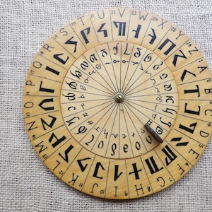 Może przedstawiać: Drewniane koło szyfrujące z dwoma obracającymi się okrągłymi dyskami. Zewnętrzny dysk zawiera litery i cyfry, a wewnętrzny symbole i cyfry. Obecny jest mały metalowy kołek i drewniany wskaźnik.