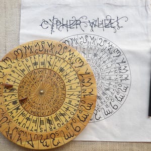 May include: A wooden cipher wheel with black markings and a brass center pin. The wheel is next to a white cloth with a printed cipher wheel design and the word "CYPHER WHEEL". A black pencil rests nearby.