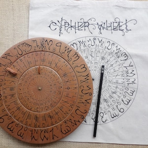 Könnte beinhalten: Ein hölzernes Chiffrier-Rad mit eingravierten Buchstaben und Symbolen, neben einer Stofftasche mit der Aufschrift "CYPHER WHEEL" und einem Chiffrier-Rad-Diagramm. Ein schwarzer Bleistift liegt auf der Tasche. Das Holzrad hat einen Durchmesser von etwa 25 cm.
