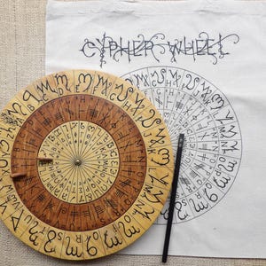Puede incluir: Una rueda de cifrado de madera con un dial interno giratorio, con símbolos y letras grabados. Un lápiz negro descansa junto a la rueda y una bolsa de mano blanca con las palabras "CYPHER WHEEL" está en el fondo. La rueda tiene unos 25 cm de diámetro.