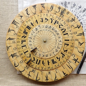 Może przedstawiać: Drewniane koło szyfrujące z koncentrycznymi obracającymi się pierścieniami, wytrawionymi symbolami i literami. Koło ma mosiężny środek i towarzyszy mu ołówek i papier z podobnymi symbolami. Tło to tkanina podobna do worka.