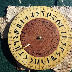 Może przedstawiać: Koło runiczne z drewna, okrągłe, z brązowym środkiem i żółtym zewnętrznym pierścieniem. Zewnętrzny pierścień ma 26 run, a wewnętrzny pierścień ma 26 liter alfabetu. Koło ma mały metalowy wskaźnik.