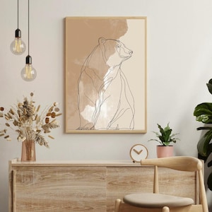Puede incluir: Una impresión artística enmarcada con un dibujo lineal minimalista de un oso sentado. La obra de arte tiene una paleta de colores neutros con tonos beige y marrón. La impresión se muestra en un marco de madera clara, agregando un toque de calidez natural a la decoración.