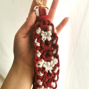 Puede incluir: Un llavero de macramé hecho a mano en rojo y blanco, con un diseño floral y un cierre dorado. El llavero mide aproximadamente 15 cm de largo, incluyendo el cierre y la borla.