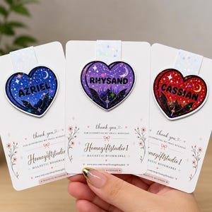May include: Three magnetic bookmarks with heart-shaped designs. Each heart features a name: Azriel, Rhysand, and Cassian, set against a starry night sky and bat silhouettes. The bookmarks are attached to white cards with floral accents and the text "thank you".