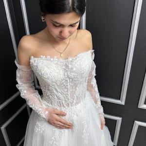 May include: A white off-the-shoulder wedding dress featuring long sheer sleeves and a bodice adorned with intricate floral lace embroidery, beadwork, and sequins. The gown has a sweetheart neckline and a full, layered tulle skirt with lace appliqués.