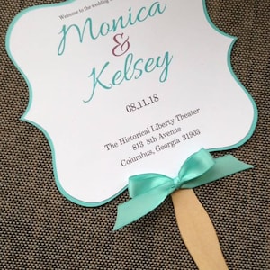 May include: Wedding ceremony program fan with a teal border and a teal ribbon. The fan features the names "Monica & Kelsey" and the date "08.11.18". The text also includes the location: "The Historical Liberty Theater, 813 8th Avenue, Columbus, Georgia 31903".