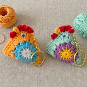 Puede incluir: Dos llaveros de crochet hechos a mano con forma de gallo. Uno es naranja con una flor azul, el otro es verde menta con una flor morada. Ambos tienen crestas rojas, detalles amarillos, ojos saltones y aros dorados.