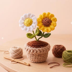 May include: A crocheted flower arrangement featuring a white daisy and a yellow sunflower in a brown and tan pot. Green leaves and a wooden crochet hook are also visible, along with several balls of yarn in various colors.