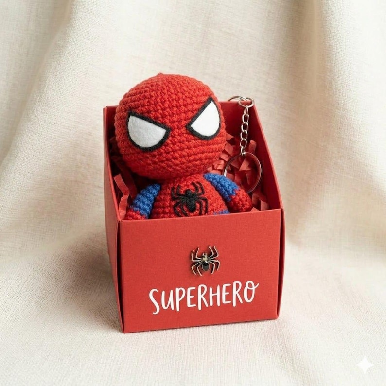 May include: A red and blue crocheted Spider-Man plush keychain with white eyes and a black spider emblem on the chest, nestled inside a red gift box featuring a small metal spider icon and the white printed text SUPERHERO on the front.