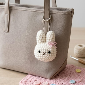 May include: A cream-colored, crocheted bunny keychain with a pink bow, black eyes, and a cross-stitched mouth hangs from a gray tote bag. The bunny is attached to a silver chain. A pink crocheted coaster and yarn are in the background.