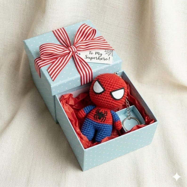 May include: A light blue gift box with white polka dots, topped with a red and white striped ribbon and a tag reading To My Superhero. Inside, nestled in red tissue paper, is a small crocheted Spider-Man figure attached to a metal keychain.