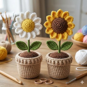 Könnte beinhalten: Zwei gehäkelte Blumenpflanzen, eine Gänseblümchen und eine Sonnenblume, in beigen Töpfen. Das Gänseblümchen hat weiße Blütenblätter und eine gelbe Mitte, die Sonnenblume hat gelbe Blütenblätter und eine braune Mitte. Beide haben grüne Blätter und Stiele.