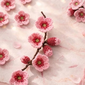 May include: Handmade crochet flowers in shades of pink and red with green centers. The flowers are arranged on a brown stem, with some individual flowers scattered around. The craftwork is delicate and detailed, perfect for spring-themed crafts.