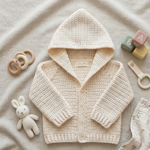 May include: A cream-coloured, hooded baby cardigan with a button closure. The crochet sweater is surrounded by baby toys, including wooden blocks, a bunny, and a teething ring. The cardigan has a tag that reads "rollingelle".