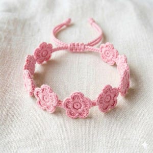Puede incluir: Una pulsera rosa de ganchillo con diseños en forma de flor. La pulsera es ajustable con un cierre de nudo corredizo. Las flores están espaciadas uniformemente, creando un accesorio delicado y femenino.