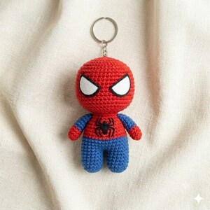 May include: A crochet keychain featuring a small, stuffed figure of a character with a red head, white triangular eyes, blue arms and legs, and a black spider emblem stitched onto the center of the red chest, attached to a silver metal ring.