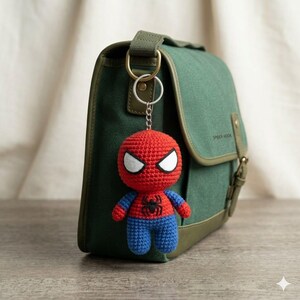 May include: A crochet Spider-Man keychain featuring a red and blue yarn figure with white eyes and a black spider emblem on the chest, attached to a silver metal ring, hanging from a dark green canvas messenger bag with a buckle closure.