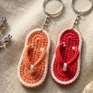 May include: Two crochet flip-flop keychains. One is orange with a white border and strap, the other is red with a white border and strap. Both have silver key rings attached. The keychains are on a beige linen surface.