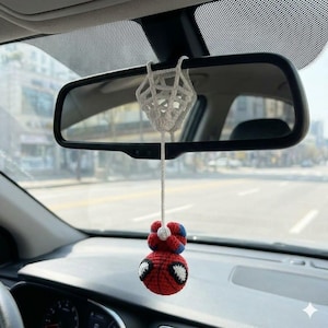 May include: A crocheted hanging ornament suspended from a rearview mirror, featuring a red and blue character head with large white eyes attached to a white crocheted web-like structure by a long white string, creating a decorative car accessory.