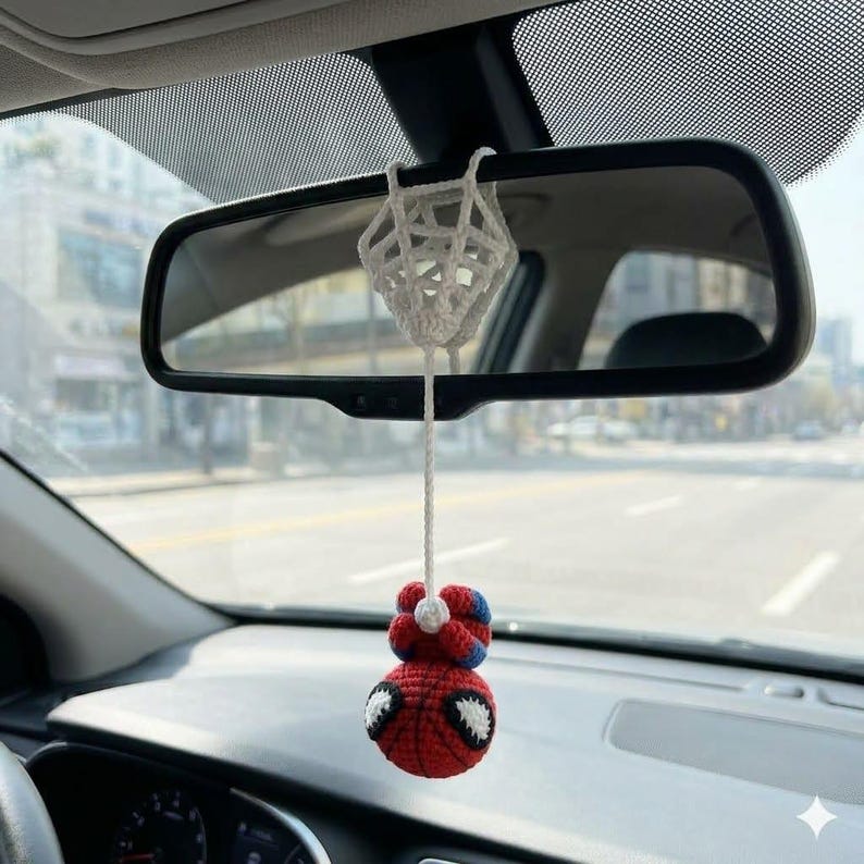 May include: A crocheted hanging ornament suspended from a rearview mirror, featuring a red and blue character head with large white eyes attached to a white crocheted web-like structure by a long white string, creating a decorative car accessory.