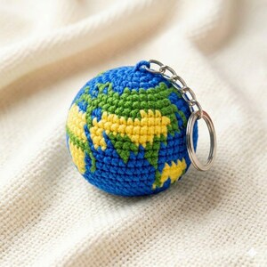 May include: A spherical crochet keychain shaped like a globe, featuring blue yarn for the oceans and yellow and green yarn for the continents. A silver-toned metal split ring is attached to the top of the globe with a short metal chain link.