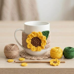 May include: A white ceramic mug with a crocheted sunflower cozy. The cozy is tan with a yellow and brown sunflower. Additional crochet items include a coaster, yarn balls, and a crochet hook. The mug is on a wooden surface.