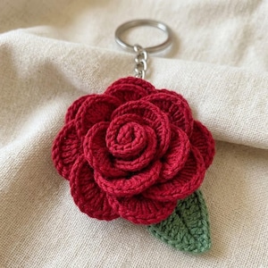 May include: A handmade red crochet rose keychain with a green leaf. The rose is detailed with multiple layers of petals and attached to a silver-colored keyring. The keychain is a charming accessory.