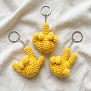 May include: Three yellow crocheted hand keychains. One keychain shows the middle finger, another shows the 'I love you' hand gesture, and the third shows the peace sign. Each keychain has a silver-colored ring.