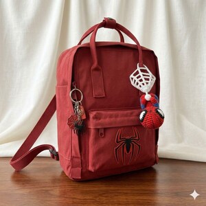 May include: A dark red backpack featuring a black spider emblem on the front pocket. The bag includes a silver keychain with a small black spider charm and a white crocheted spiderweb accessory with a red and blue crocheted spider figure.