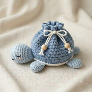May include: A light blue crochet turtle drawstring pouch featuring a rounded shell body, a small head with embroidered closed eyes, and four short flippers. The pouch is cinched with a white cord tied in a bow, accented by two wooden beads.