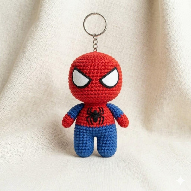 May include: A crocheted Spider-Man keychain featuring a round red head with large white eyes, a red torso with a black spider emblem, and blue arms and legs. The figure is attached to a silver-colored metal keyring with a short chain link.