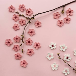 以下が含まれることがあります： ピンクと白のかぎ針編みの花のアップリケがピンクの背景に散らばり、茶色の枝があります。花の中心は濃い赤色です。アップリケはさまざまなサイズで、花の模様のように配置されています。