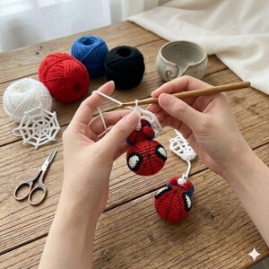 May include: A crochet project featuring two small red and blue spherical figures with black web patterns and white eyes, being worked on with a wooden crochet hook. Nearby are balls of white, red, blue, and black yarn, a white web motif, and scissors.