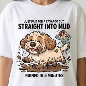 Puede incluir: Camiseta blanca con una ilustración de dibujos animados de un perro Cavapoo embarrado. El texto dice: "JUST PAID FOR A CAVAPOO CUT STRAIGHT INTO MUD RUINED IN 5 MINUTES". Una persona se muestra en el fondo.