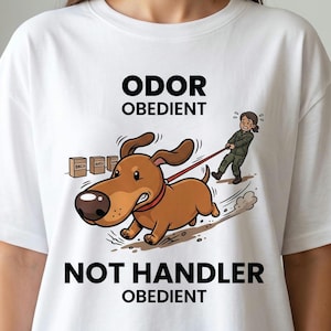 May include: White t-shirt with a humorous cartoon dog illustration. The dog, wearing a red collar, is pulling a handler, with the text "ODOR OBEDIENT" and "NOT HANDLER OBEDIENT" above and below the image.