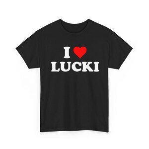 Könnte beinhalten: Ein schwarzes Kurzarm-T-Shirt mit dem weißen Text I, gefolgt von einem roten Herzsymbol und dem Wort LUCKI in fetten, weißen Großbuchstaben, mittig auf der Brust. Das Kleidungsstück hat einen Rundhalsausschnitt und einen geraden Schnitt.