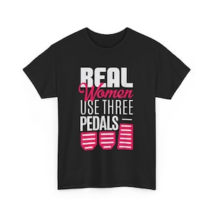 Puede incluir: Una camiseta negra de manga corta con texto en blanco y rosa que dice REAL Women USE THREE PEDALS. Debajo del texto hay tres iconos gráficos de color rosa que representan pedales de coche, siendo el pedal derecho más alto que los otros dos.
