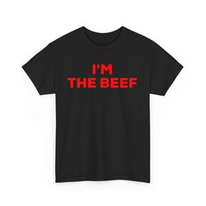 Op de afbeelding: Een zwart T-shirt met korte mouwen en de tekst I'M THE BEEF in vetgedrukte, felrode hoofdletters in het midden op de borst. Het kledingstuk heeft een standaard ronde hals en een rechte zoom, gepresenteerd tegen een effen achtergrond.
