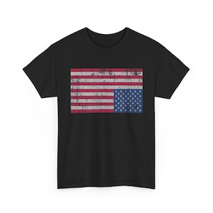 Puede incluir: Una camiseta negra de manga corta con un gráfico rectangular en el pecho. El gráfico muestra una bandera de los Estados Unidos con un estilo desgastado, con rayas horizontales rojas y blancas y un campo azul con estrellas blancas a la derecha.
